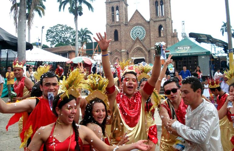 Un gruppo di uomini e donne festeggia in strada durante il Festival del Soltero (o Festa dei single) a Chaguaní, in Colombia. Indossano costumi colorati rosso e oro, con corone e copricapi decorati con spighe dorate. Alcuni lanciano coriandoli e sorridono verso la fotocamera, creando un’atmosfera vivace e allegra. Sullo sfondo si vede la piazza del paese con una chiesa di mattoni e palme alte, oltre a bancarelle e persone che osservano la sfilata.